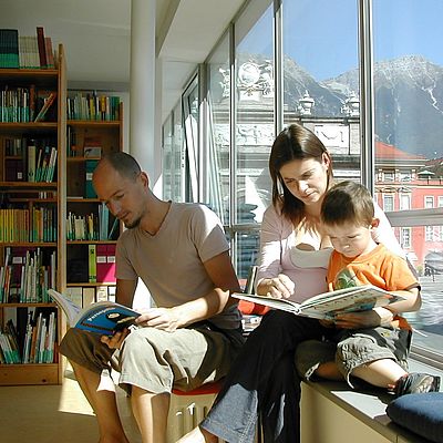 Bücherregal in der Nord-Süd-Bibliothek Innsbruck. Ein Mann liest, daneben blättert eine Frau mit einem Kind durch ein anderes Buch