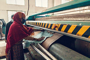 In Sari gekleidete Frau mit Mund-Nasenschutz betätigt eine große Maschine in einer Textilfabrik in Bangladesch für Lederwaren und Schuhe