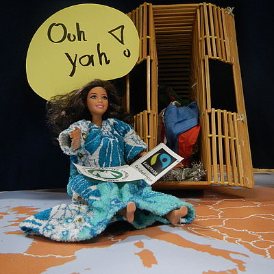 Barbiepuppe in buntem Kleid sitzt auf einer Weltkarte. Baumwoll Mode Workshop