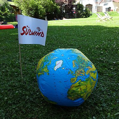 Aufgeblasener Wasserball bedruckt mit einer Weltkarte, daneben steckt im Boden eine Südwind Fahne 