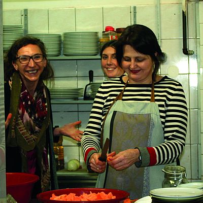 Drei Frauen bereiten in einer Küche Essen vor, lachen dabei in die Kamera