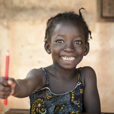 Ein lachendes Mädchen streckt ihren Arm in Richtung Kamera aus. In ihrer Hand ist ein roter Stift