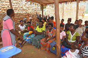 Eine Frau der Village Health Teams in Uganda hält einen Vortrag vor einer Menschenmenge unter einem Holzverschlag