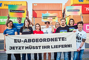 Aktivist:innen der Kampagne "Menschenrechte brauchen Gesetze" stehen vor dem Containerhafen in Wien und halten ein Transparent mit der Aufschrift: "EU-Abgeordnete jetzt müsst ihr liefern"