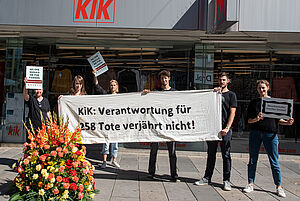 Sechs in schwarz gekleidete Aktivistinnen und Aktivisten halten Banner und Plakate vor einer KiK-Filiale auf denen steht: "Kik: Verantwortung verjährt nicht!"
