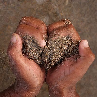 Zwei Hände formen ein Herz und halten dabei Sand. Im Hintergrund Meer und Sand