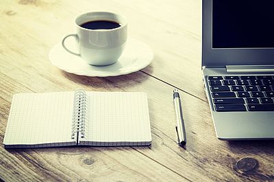 Neben einem aufgeklappten Laptop liegt ein Stift, ein offener Notizblock und eine Tasse Kaffee