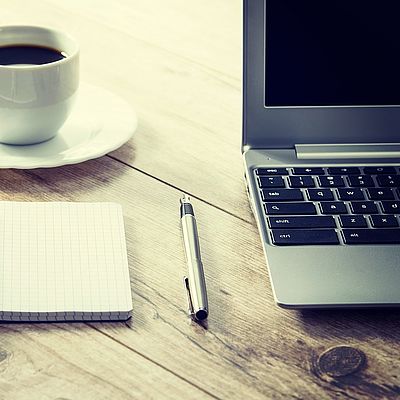 Neben einem aufgeklappten Laptop liegt ein Stift, ein offener Notizblock und eine Tasse Kaffee