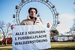 Ein junger Mann steht vor dem Wiener Riesenrad mit einem Protestschild für Waldschutz