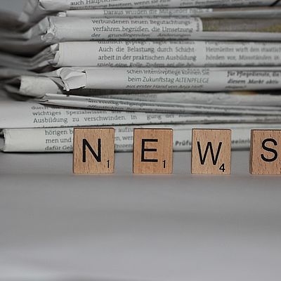 Vier Scrabble-Steine bilden das Wort "News". Im Hintergrund sind mehrere Zeitungen gestapelt