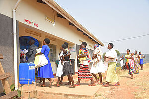 Eine Schlange an afrikanischen Frauen, viele mit Babys im Arm, wartet in der Sonne vor dem Eingang der Entbindungsstation des Maisuka-Krankenhauses