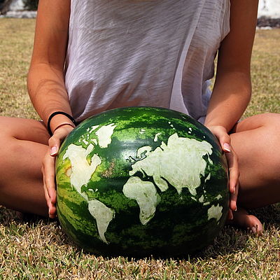 Zwei Hände halten eine Wassermelone. Auf ihrer Oberfläche ist die Weltkarte eingeritzt.