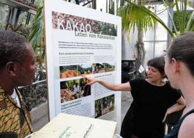 Zwei Personen betrachten ein Ausstellungsplakat, eine dritte Person deutet mit dem Finger auf das Plakat