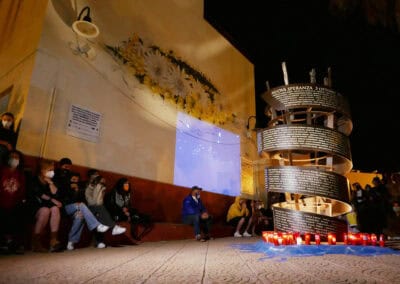 Menschen sitzen bei einer nächtlichen Zeremonie um ein spiralförmiges Mahnmal mit Namen von Opfern der Flucht über das Mittelmeer; Kerzen stehen am Boden.