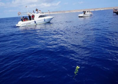 Menschen auf mehreren Booten auf offenem Meer, eine einzelne weiße Blume treibt im Wasser – Symbol der Erinnerung an verstorbene Flüchtlinge.
