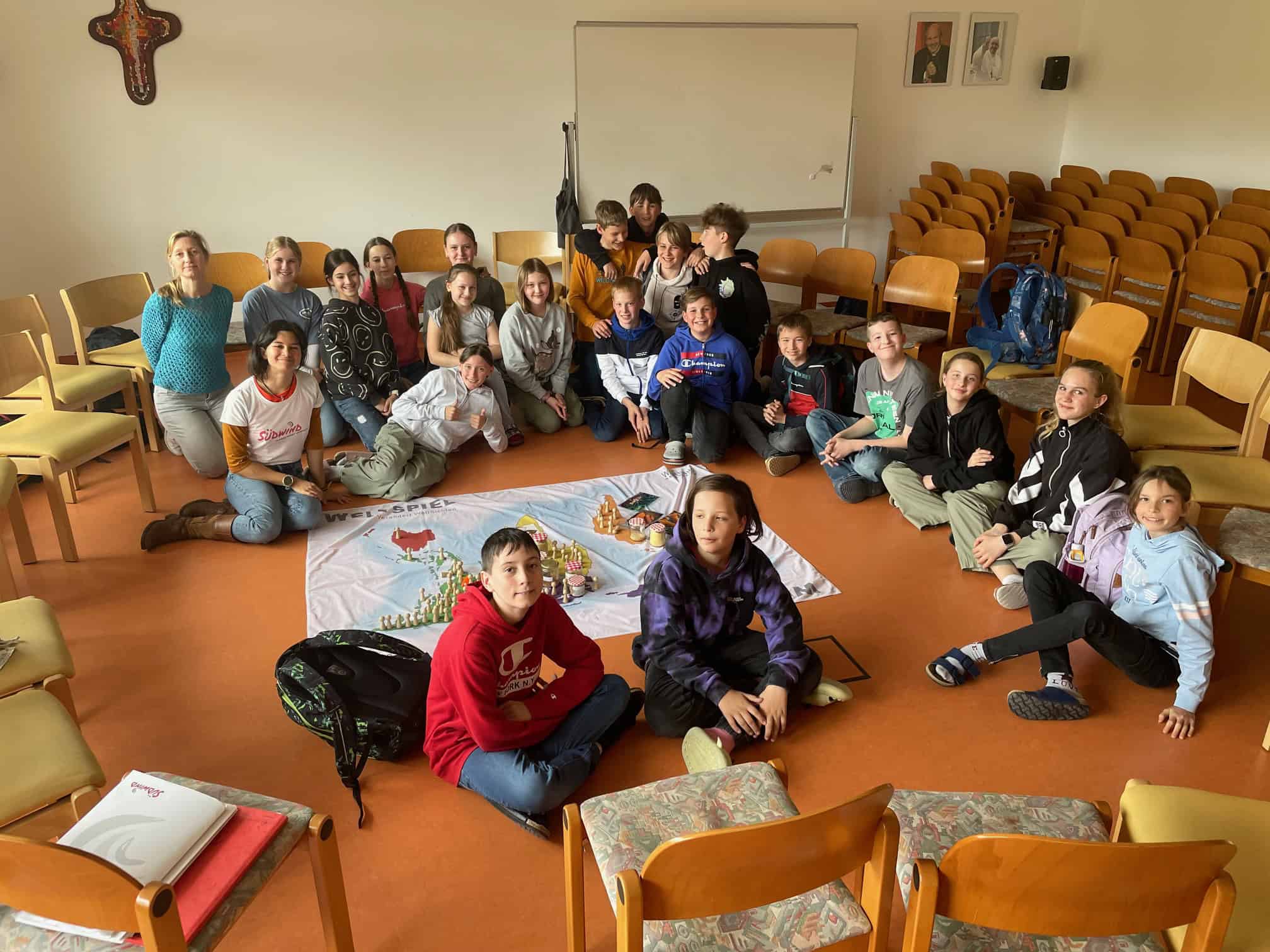 LEADER Generationen FAIRbinden Workshoptag Suedwind: Kinder und Jugendliche sitzen in einem Kreis rund um ein ausgebreitetes Weltspiel in einem Raum mit Holzstühlen. Alle wirken aufmerksam und engagiert.