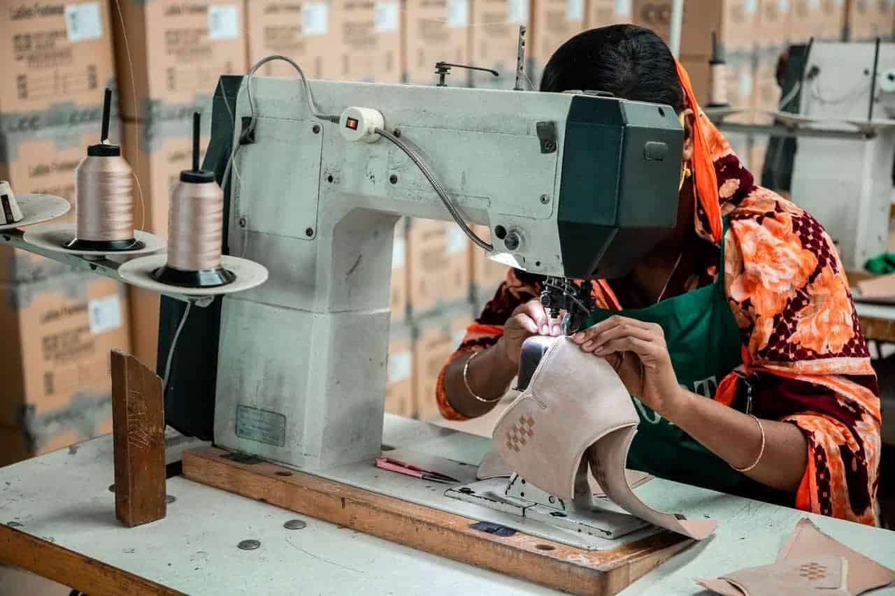 Frau in farbenfroher Kleidung näht konzentriert ein Lederteil an einer großen Industriemaschine in einer Fabrik mit Kartonstapeln im Hintergrund.