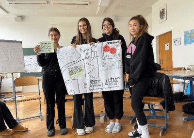 Vier Schülerinnen halten ein großformatiges Plakat mit Illustrationen zum Thema Nachhaltigkeit hoch. Sie stehen vor einer Pinnwand und einem Flipchart.