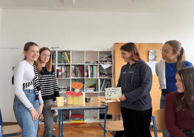 Fünf Schülerinnen präsentieren in einem Klassenzimmer ein selbstgebautes Modell und beschriftete Karten mit Botschaften zu Nachhaltigkeit. Zwei sitzen im Vordergrund, drei stehen um den Tisch herum.