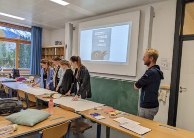 Fünf Schüler:innen stehen an einem Tisch und arbeiten an einem großen Papierbogen. Ein Lehrer schaut zu. Im Hintergrund ist ein Beamerbild mit Text zur kritischen Mediennutzung zu sehen.