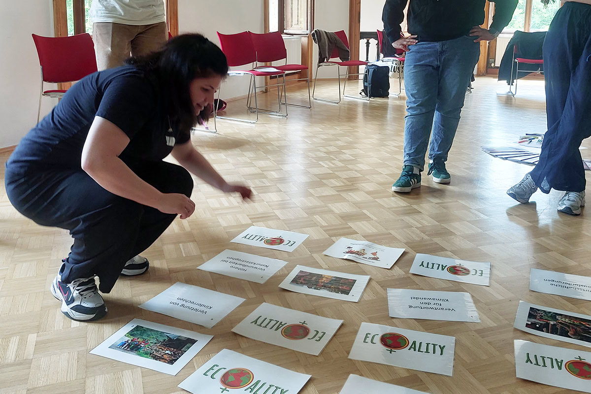 2025 05 Teilnehmer:innen des Kochworkshops für klimagerechte Ernährung bei der Südwind Academy im Mai 2025 in Wels(c) Suedwind (10) Eine Frau hockt sich in einem Seminarraum über mehrere auf dem Boden verteilte DIN-A4-Blätter mit Texten und Bildern. Weitere Personen stehen um sie herum und betrachten die Inhalte.