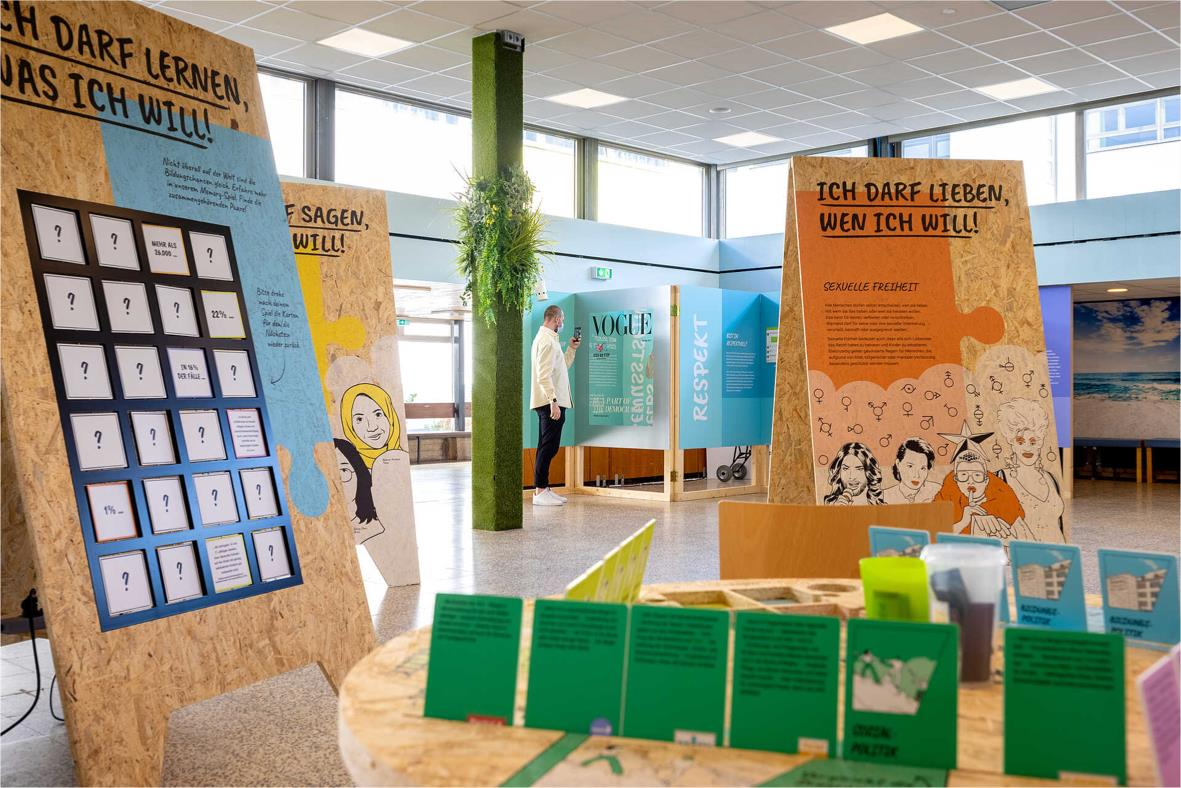 Step by Step Demokratieräume – Ausstellungsflyer-1-5 Das Bild zeigt in der Mitte einen Tisch mit Kärchen und im Hintergrund Ausstellungsexponate.