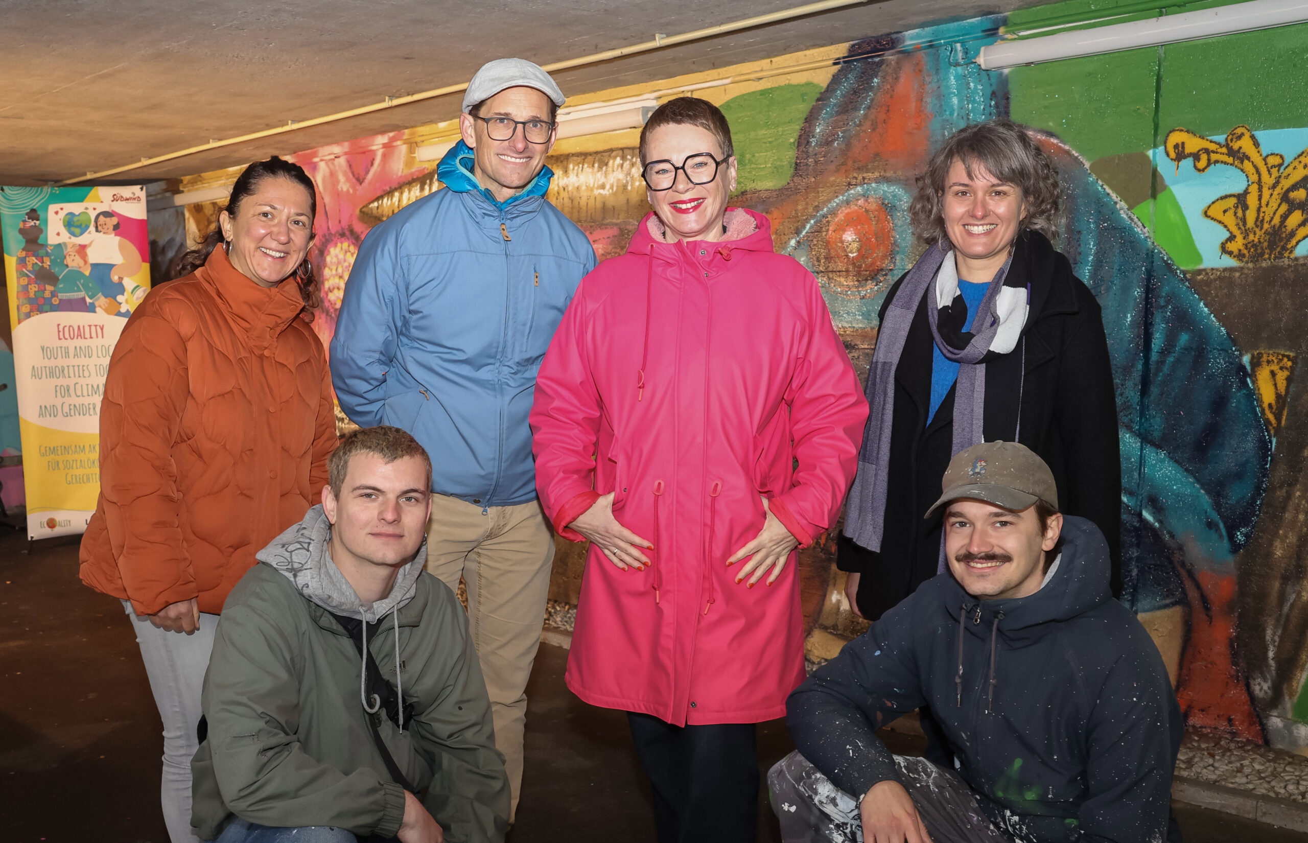 20251111d_druck Lisa Aigelsperger (Südwind), David Eichinger (Ramsauer Gymnasium), Stadträtin Mag.a Eva Schobesberger und Anna Riebe-Maybach (Ramsauer Gymnasium) (von links nach rechts) Moritz Danner und Mario Moder von der Kunstuni Linz (vorne) vor dem Mural des Ramsauergymnasium