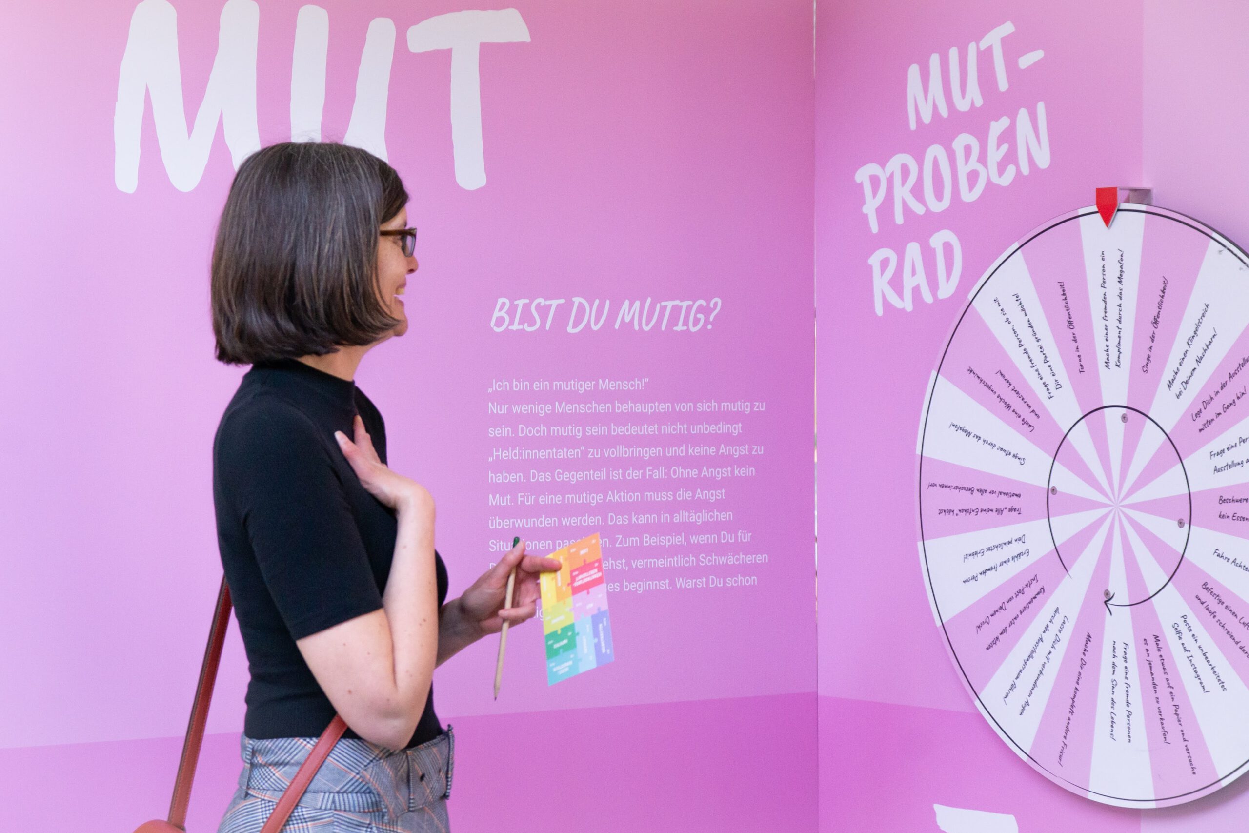 StaBiGröpelingen_(c) Jennifer Pfeifer, Step by Step Eine Frau steht vor der Station der Ausstellung mit dem Mutprobenrad und lacht bestärkt.