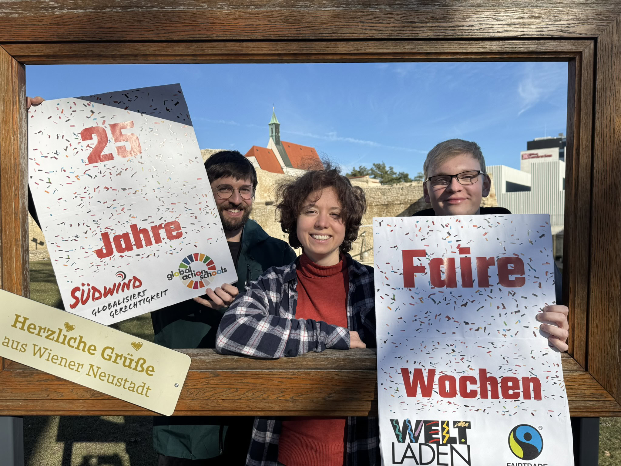 Faire Wochen_c_Südwind Zwei Männer und eine Frau halten gut gelaunt bei Sonnenschein im Freien Schilder in die Höhe.