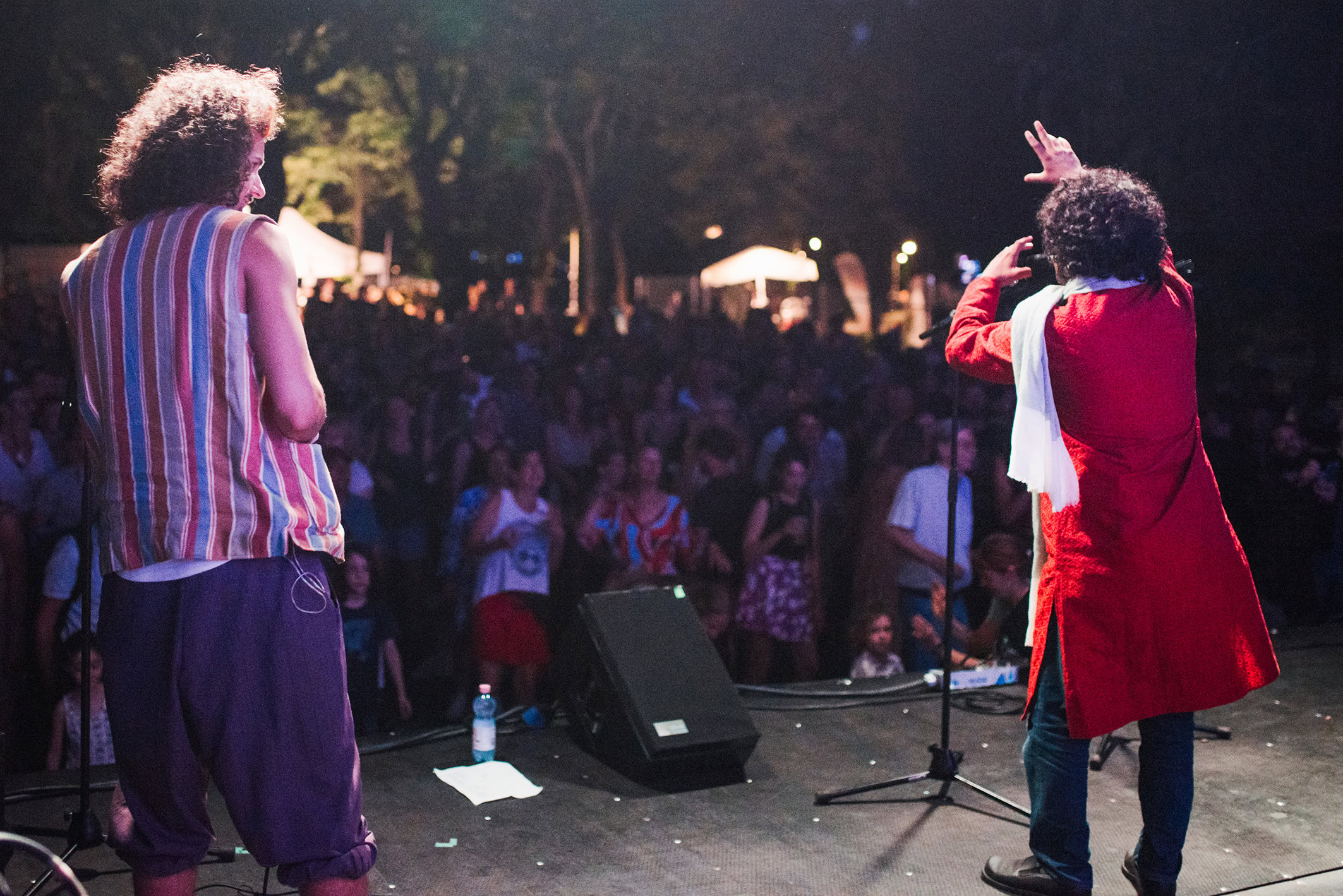 Zwei Männer stehen auf einer abgedunkelten Bühne und performen. Der Mann links trägt ein gestreiftes Hemd und eine dunkle Hose, der Mann rechts ein rotes Gewand mit einem Schal. Im Hintergrund ist ein Publikum zu erkennen, das die Performance verfolgt. Die Atmosphäre ist intensiv und emotional.