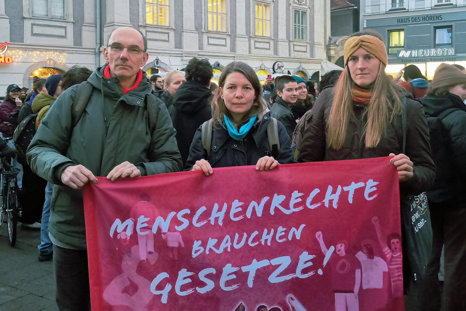 3 Personen bei einer Kundgebung mit einem Banner: "Menschenrechte brauchen Gesetze!"