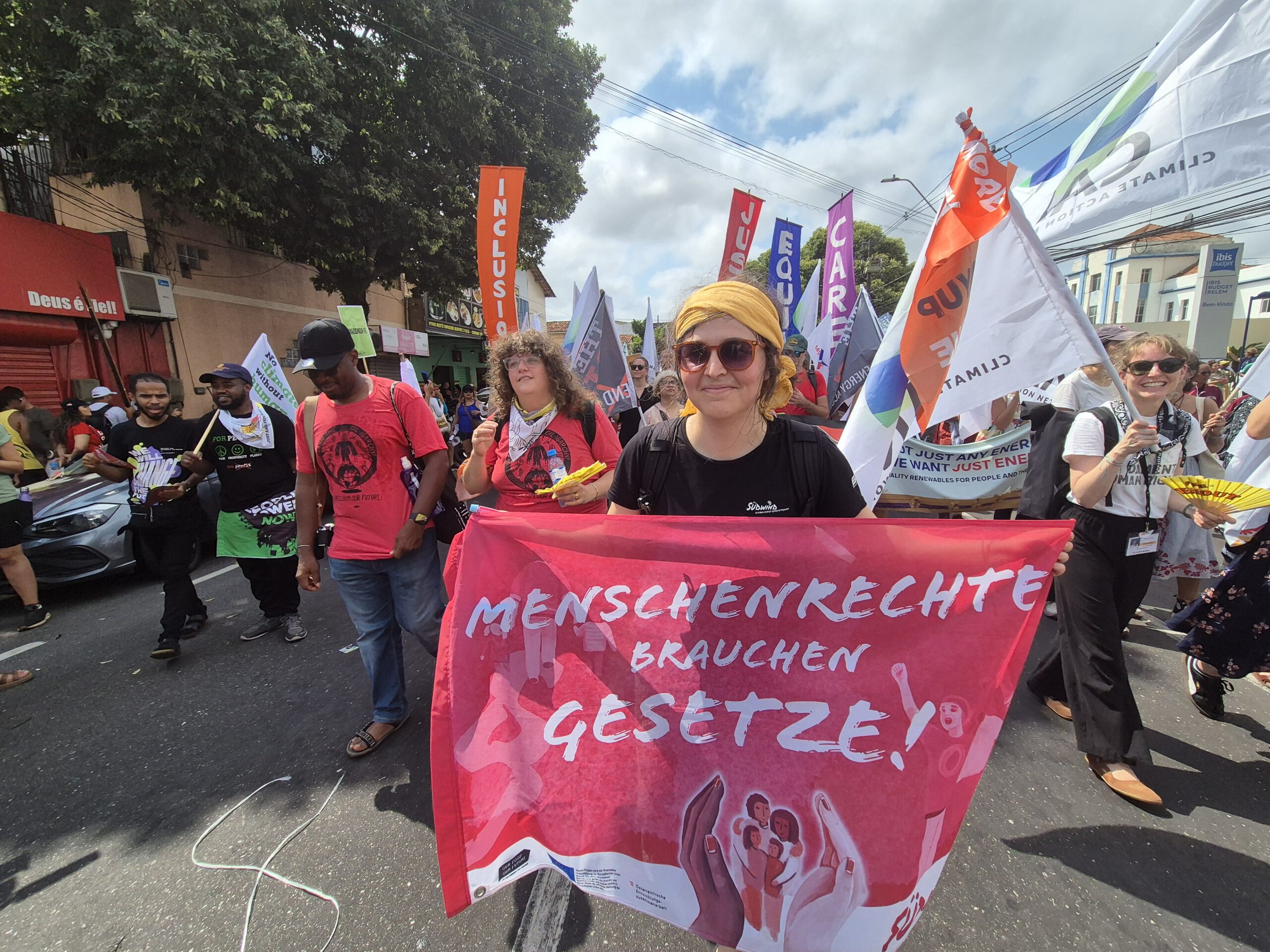 Gruppe Demonstrant:innen in Belem, Brasilien im Zuge der Welklimkonferenz COP30. Sie tragen bunte Flaggen und Schilder mit Forderungen für Klimagerechtigkeit und soziale Gerechtigkeit. Im Vordergrund hält Südwind-Sprecherin Angelika Derfler ein Transparent mit der Aufschrift "MENSCHENRECHTE BRAUCHEN GESETZE!".