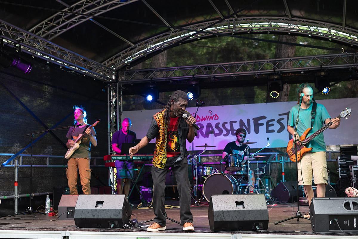 Eine Band mit fünf Musikern performt auf einer Freilichtbühne beim Südwind Straßenfest. Im Vordergrund steht ein Sänger mit Dreadlocks und buntem Oberteil, der ein Mikrofon hält. Links von ihm spielt ein Mann mit blauer Jacke E-Gitarre, rechts ein Mann mit grünem T-Shirt Bass. Im Hintergrund sind Schlagzeug, Keyboard und ein Banner mit der Aufschrift 'Südwind Straßenfest' zu sehen. Die Bühne ist von Bäumen umgeben, und die Stimmung ist energiegeladen