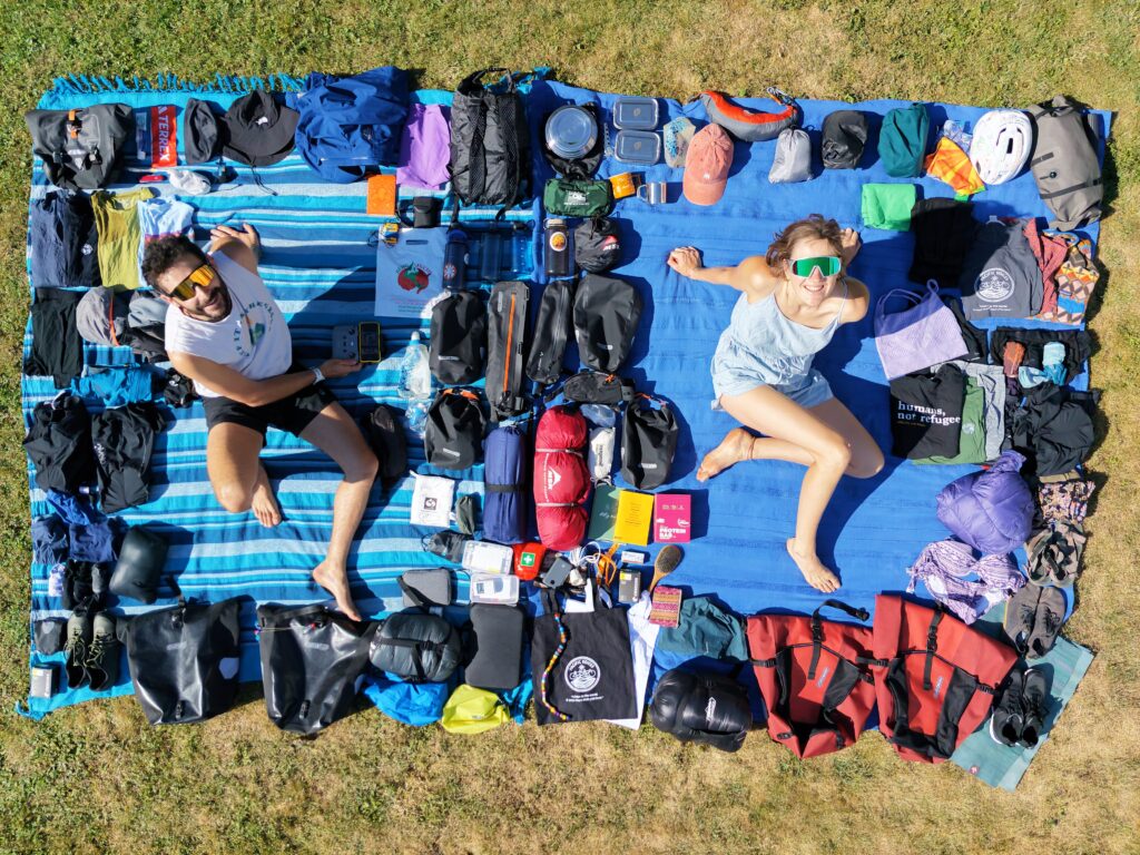Zwei Personen liegen auf einer blauen Decke auf dem Rasen, umgeben von einer großen Auswahl an Outdoor-Ausrüstung. Links liegt ein Mann mit Sonnenbrille und weißem T-Shirt, rechts eine Frau mit grünen Haarbändern und einem grauen Top. Zwischen ihnen sind Rucksäcke, Kleidung, Kochutensilien, ein Zelt und weitere Campingausrüstung ordentlich arrangiert. Die Szene wirkt abenteuerlustig und vorbereitend für eine Reise.