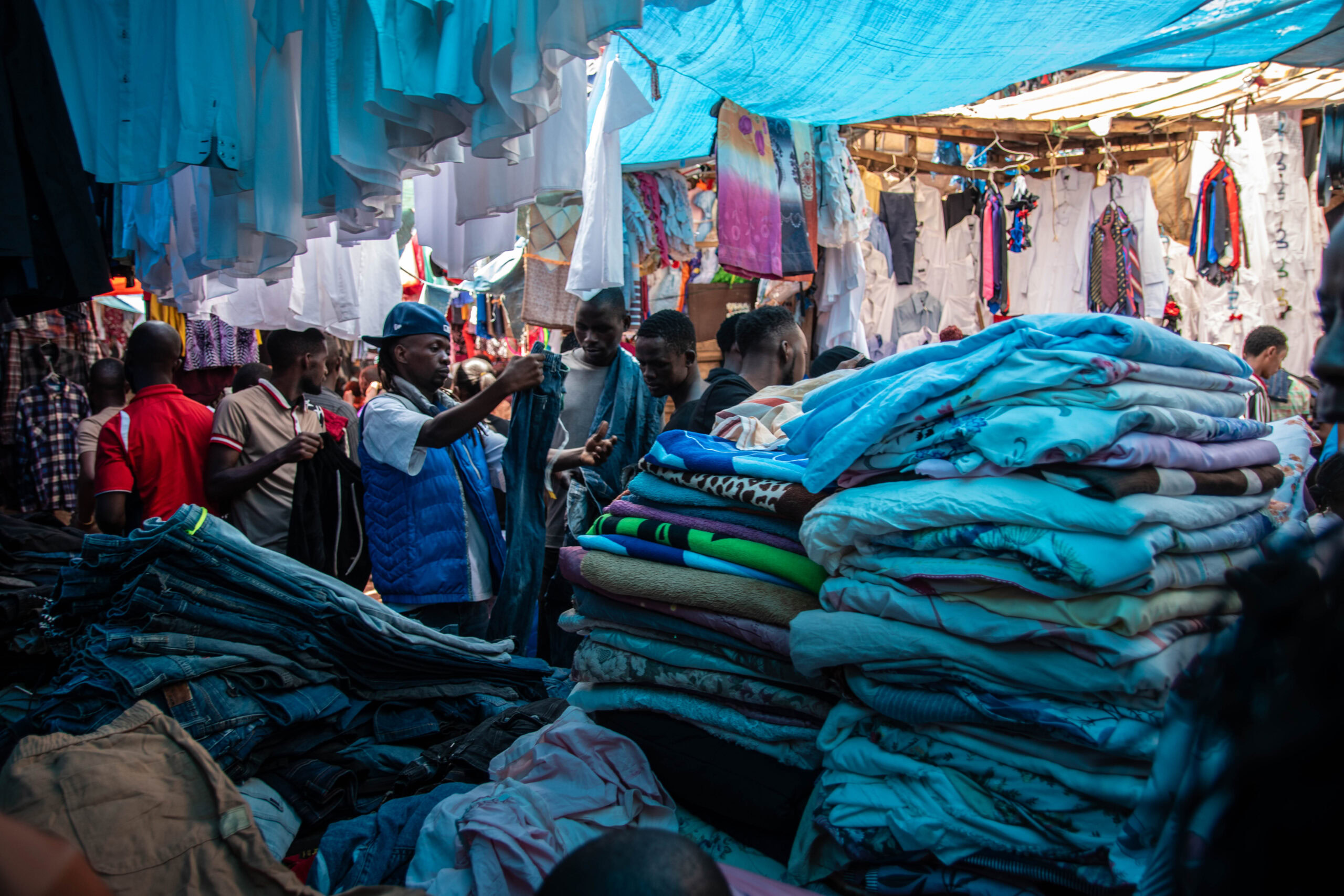 Der Owino Markt in Kampala, Uganda - Ein belebter Stoffmarkt unter einem blauen Sonnenschutz. Mehrere Männer durchstöbern Stapel von gefalteten Stoffen in verschiedenen Blautönen, die auf einem Tisch aufgetürmt sind. Im Hintergrund hängen weitere Kleidungsstücke wie Hemden und Hosen an einer Leine. Die Atmosphäre ist lebhaft und geschäftig, mit vielen Menschen, die sich um die Waren kümmern.