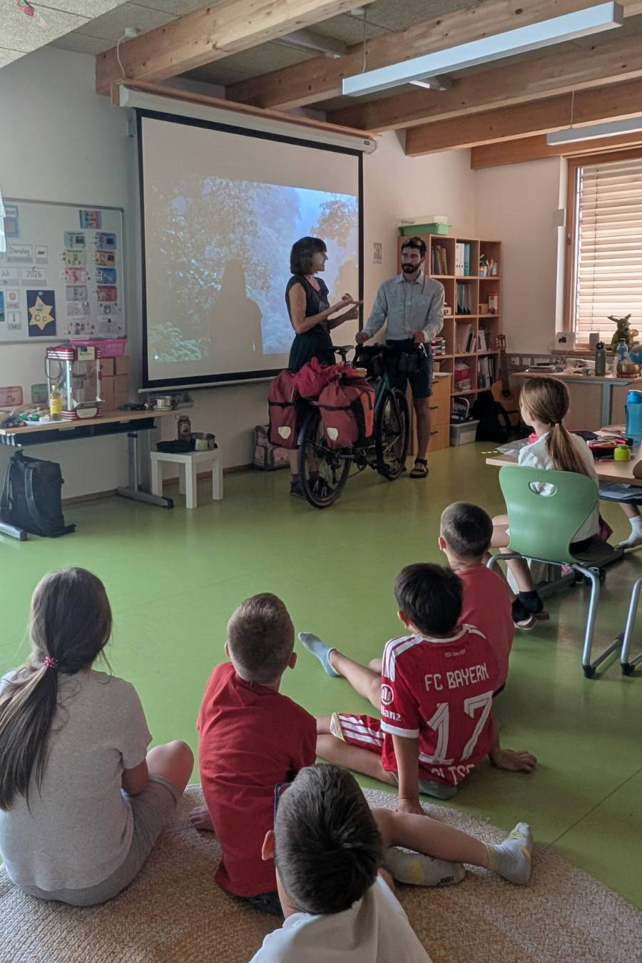 Eine Gruppe von Kindern sitzt auf dem Boden und an Tischen in einem Klassenzimmer mit Holzdeckenbalken. Vorne steht eine Frau und hält ein Mikrofon, während sie zu den Kindern spricht. Neben ihr steht ein Mann mit einem Fahrrad, das mit roten Packtaschen beladen ist. Auf der Leinwand hinter ihnen ist eine Weltkarte mit der Überschrift 'Wo geht’s lang?' zu sehen. Die Atmosphäre ist interaktiv und lehrreich.