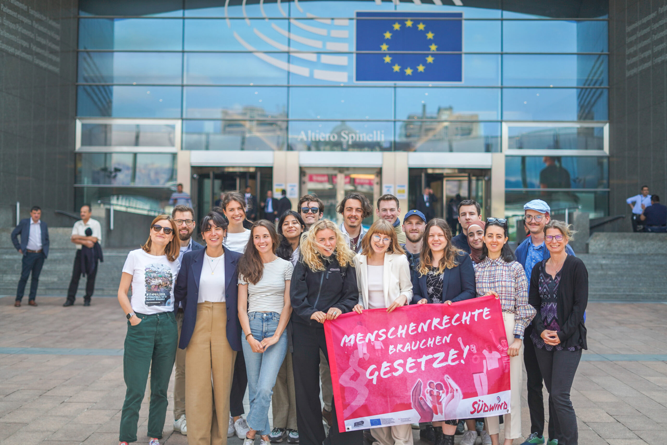 Eine Gruppe von jungen Menschen steht vor dem Eingang des Europäischen Parlaments in Brüssel. Die Gruppe hält ein rotes Banner mit der Aufschrift 'MENSCHENRECHTE BRAUCHEN GESETZE!' und dem Logo von SÜDWIND. Die Personen sind unterschiedlich gekleidet, einige tragen Anzüge oder Blazer, andere lässige Kleidung. Die Szene wirkt engagiert und politisch aktiv.