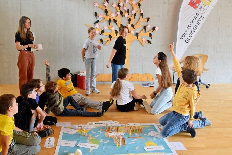 Eine Gruppe von Kindern sitzt im Kreis auf dem Boden in einem Klassenzimmer der Volksschule Gloggnitz. Im Mittelpunkt liegt eine große Weltkarte mit Kontinenten und Beschriftungen wie 'Europa', 'Nordamerika', 'Südamerika', 'Afrika', 'Asien', 'Australien' und 'Antarktis'. Eine Frau in braunen Hosen und ein Mann in einem grauen T-Shirt mit der Aufschrift 'Pacific Voices' stehen und interagieren mit den Kindern. An der Wand hängt eine Holzskulptur eines Baumes mit Vögeln. Die Szene wirkt lehrreich und interaktiv.