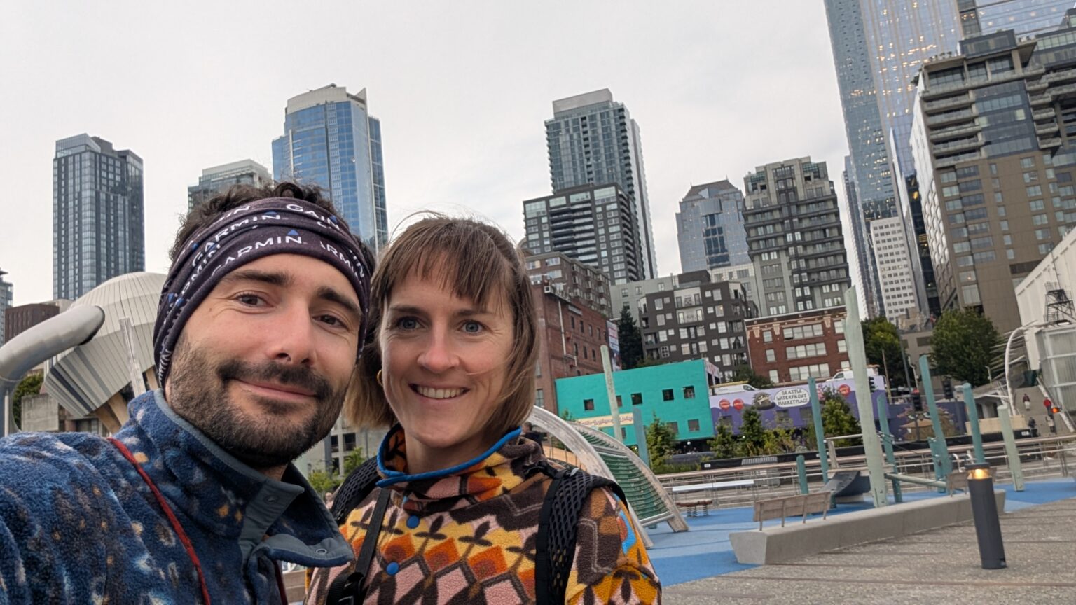 Zwei lächelnde Personen (Paul & Alina) in Fleecejacken machen ein Selfie am Waterfront von Seattle, im Hintergrund die Skyline der Stadt.