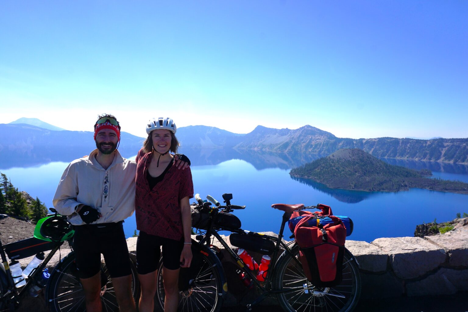 Alina und Paul mit vollbepackten Tourenbikes posieren lächelnd am Aussichtspunkt über dem Crater Lake in Oregon, dessen tiefblaues Wasser und die Wizard Island im Hintergrund zu sehen sind.